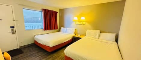 A hotel room with two beds, a window with curtains, a bedside table with a lamp, and a door.