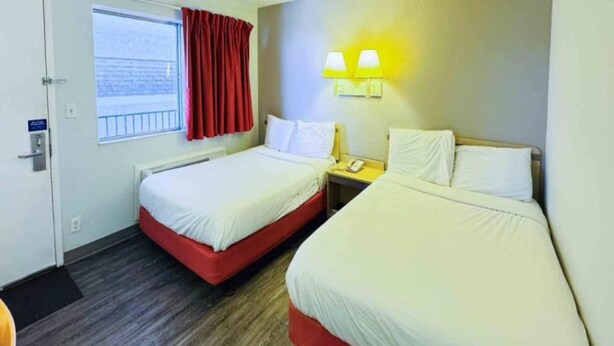 A hotel room with two beds, a window with red curtains, a bedside table with a lamp, and a door with a sign. - 2 Full Beds|Suite,Non-Smoking,Kitchen (Casper WY)