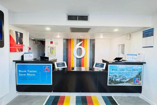 Een hotel receptie met een groot nummer 6 op de muur, twee receptiebalies en promotieposters voor een boekingsapp.