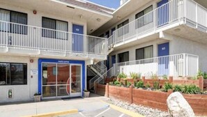 Um motel com vários andares, entrada azul, estacionamento externo e área ajardinada com plantas.