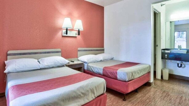 Habitación de hotel con dos camas, una mesita de noche, una lámpara de pared y un baño con lavamanos y espejo.