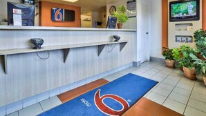 Zona de recepción de hotel con mostrador, reloj, televisión y alfombra azul con un logotipo.