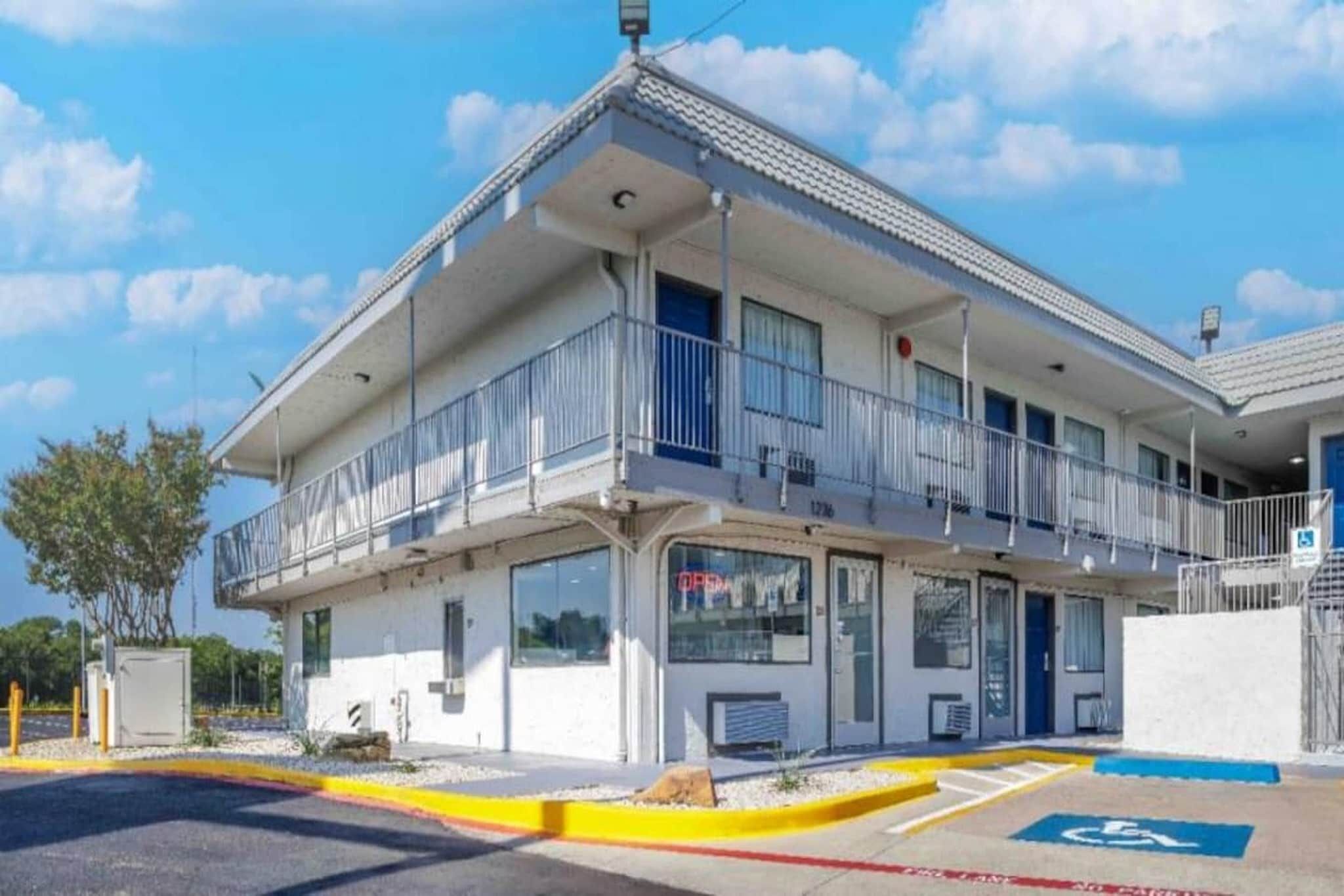A two-story motel with a balcony on the second floor, a parking lot with a handicapped parking space, and a clear blue sky.