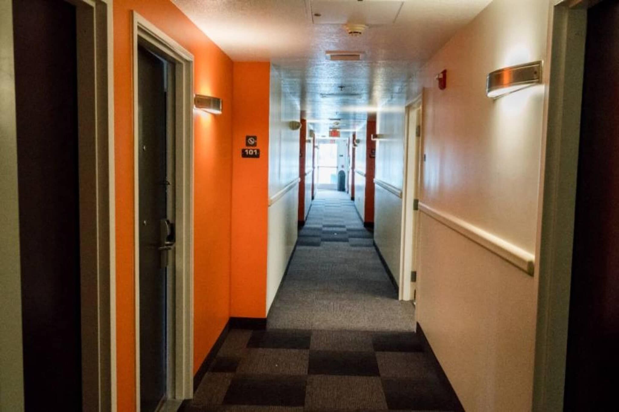 A hotel corridor with a checkered carpet, orange walls, and doors with signs.
