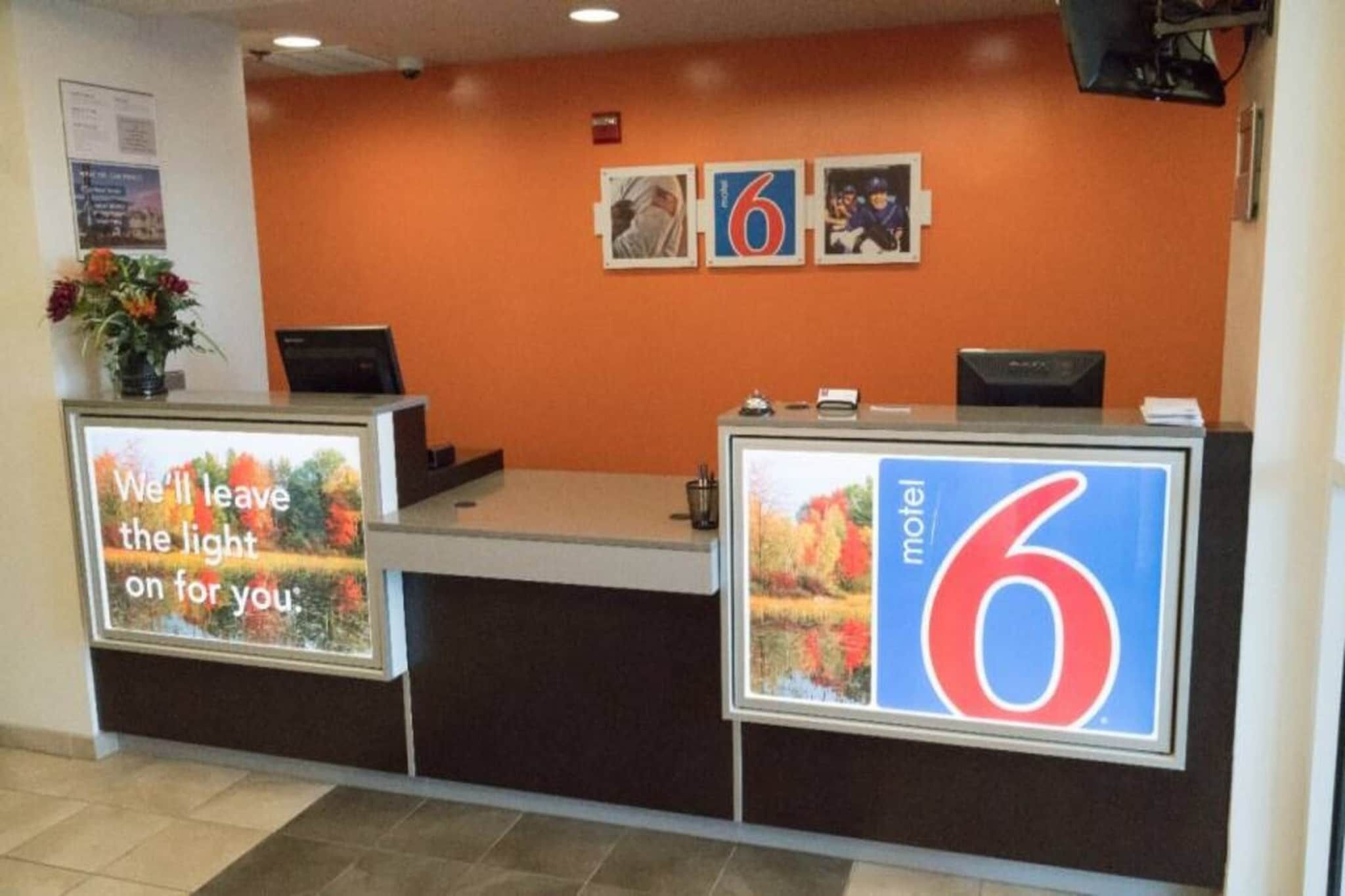 A hotel reception desk with a large sign indicating it's a '6' motel. There are two monitors, a plant, and a framed picture on the wall.
