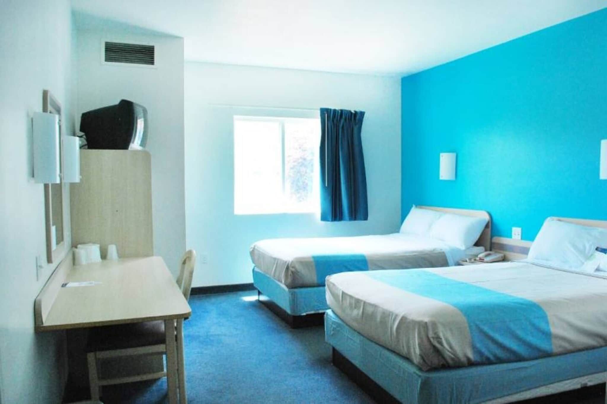 Une chambre d’hôtel avec deux lits, un bureau, une chaise, une fenêtre avec des rideaux et un mur bleu.