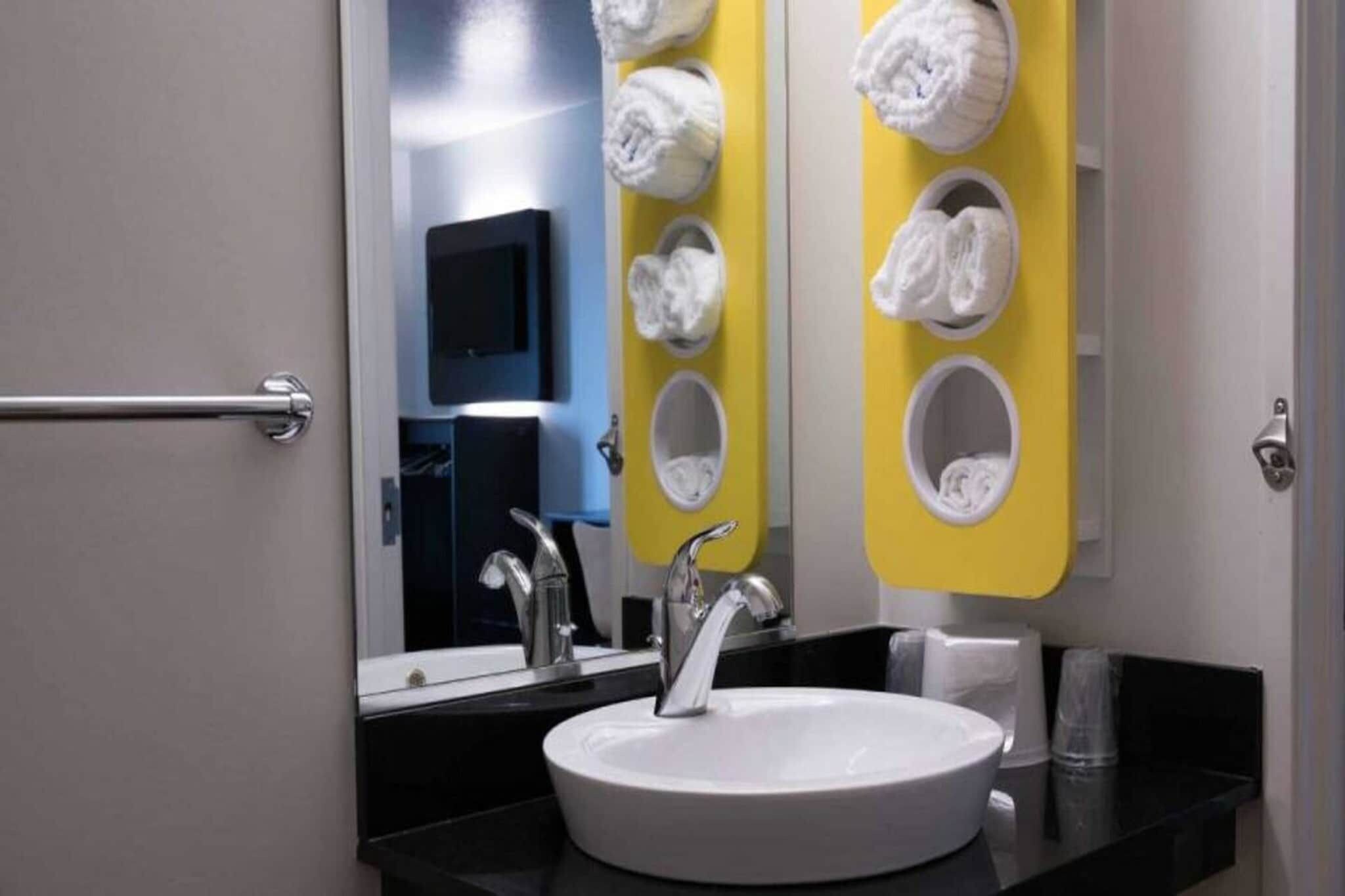 A bathroom with a sink, mirror, towel hooks, and a towel dispenser.