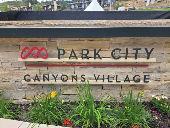 Skilt for Park City Canyons Village med en stenvæg i baggrunden og grønne planter foran.