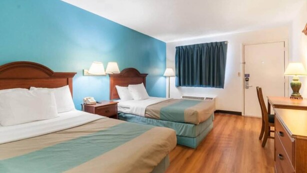 Una camera d'albergo con due letti, una scrivania, una sedia, un telefono e una finestra con tende.
