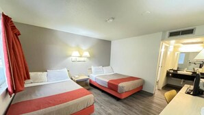 Quarto de hotel com duas camas, uma mesa de escritório, uma cadeira, uma luminária e banheiro com espelho.