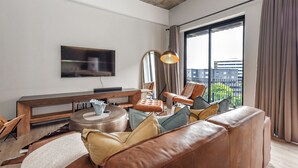 Una sala moderna con sofá de cuero, una mesa redonda de centro, un televisor de pantalla plana montado en la pared y un ventanal con cortinas.