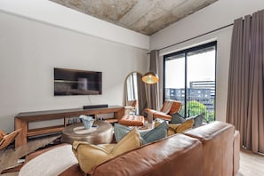 Uma sala de estar moderna com um sofá de couro, uma mesa de centro redonda, uma TV de tela plana na parede e uma janela grande com cortinas.