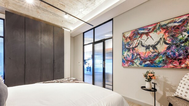 Un dormitorio moderno con una cama grande, una puerta corrediza de vidrio que da a una cocina y un cuadro abstracto y colorido en la pared.