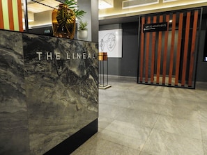 Balcão de recepção com a sinalização "THE LINEAL", uma planta em vaso e um aviso com a inscrição "LIFT TO APARTMENTS".