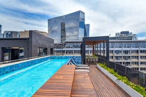 Piscina en la azotea con plataforma de madera, rodeada de edificios modernos.