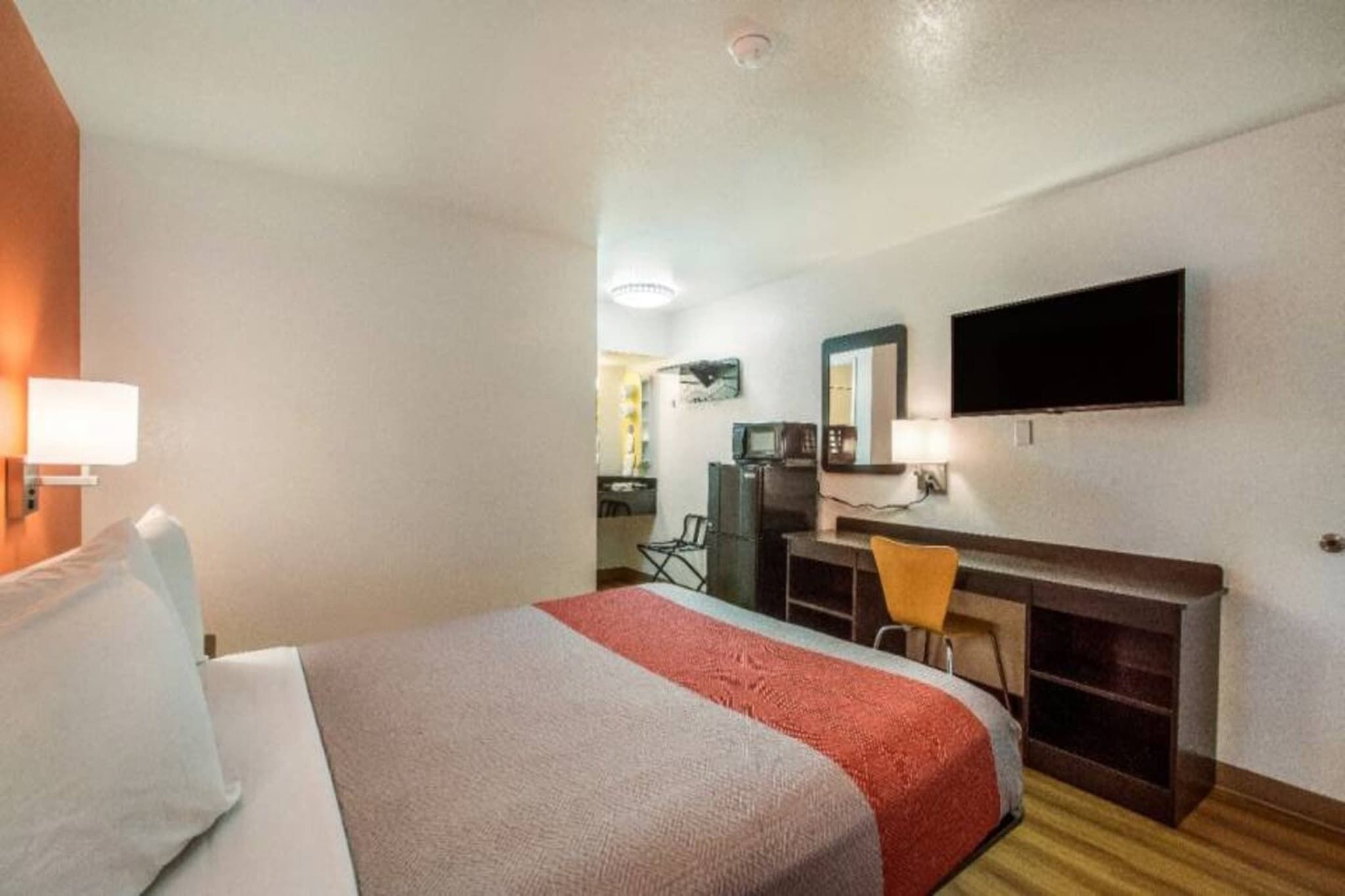 Habitación de hotel con cama, escritorio, silla, televisor y espejo.