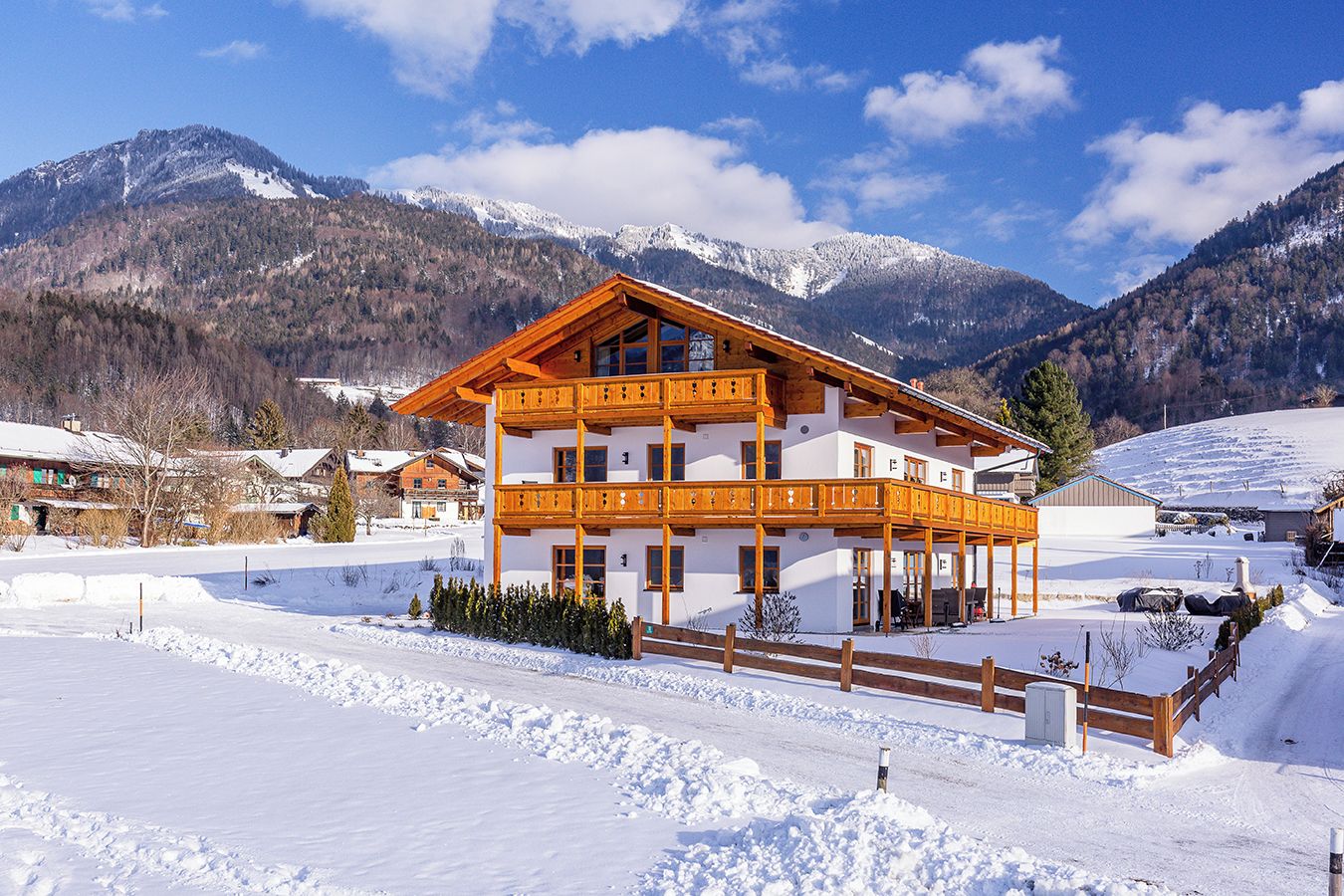 A two-story wooden chalet with balconies, surrounded by snow-covered mountains and a clear sky.
