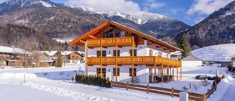 A two-story wooden chalet with balconies, surrounded by snow-covered mountains and a clear sky.