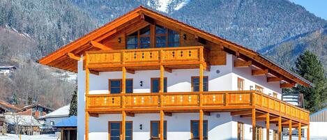 A multi-story wooden chalet with balconies, set against a snowy mountain backdrop.