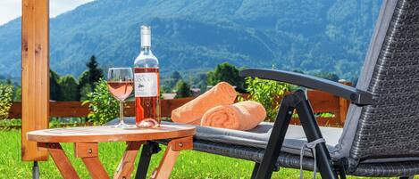 A wooden table with a bottle of wine and a glass, a wicker chair, and a towel on a patio with a mountain view.