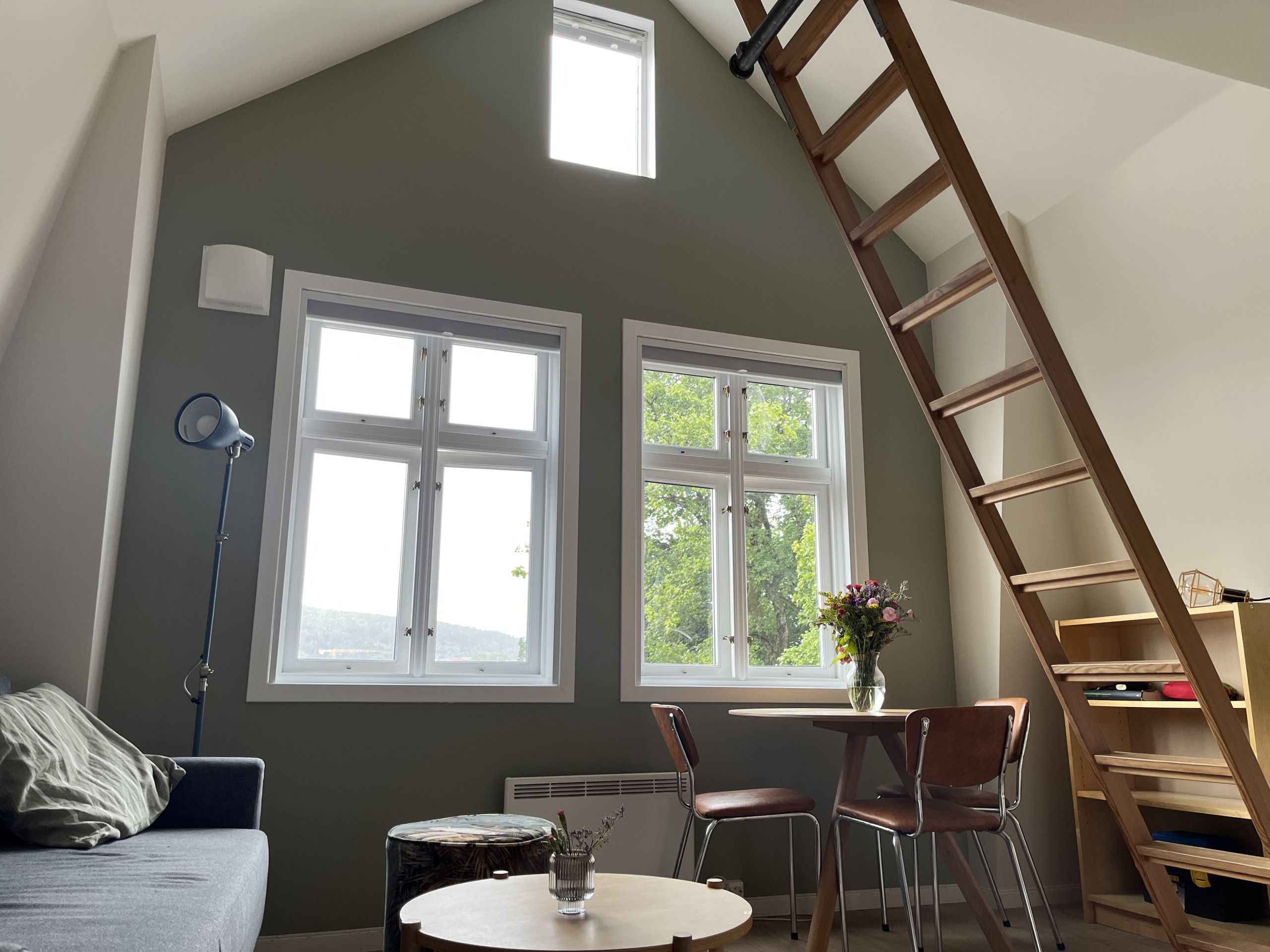 Ein Zimmer mit schrägverdeckter Decke, zwei Fenstern, einem Tisch mit Stühlen, einer Stehlampe und einer Holzleiter.