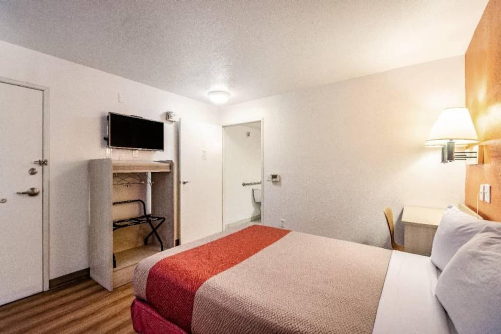 Et hotelværelse med seng, fjernsyn ophængt på væggen, skrivebord med lampe og et klædeskab.