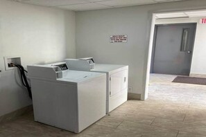 Een wasserette met twee witte wasmachines en drogers, een bord aan de muur en een deur naar een andere ruimte.