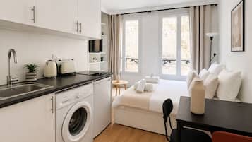 A compact hotel room with a kitchenette, a washing machine, a bed with pillows, a small table, and a chair.