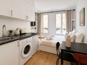 Une chambre d’hôtel compacte comprenant un coin cuisine, un lave-linge, un lit avec des oreillers, une petite table et une chaise.