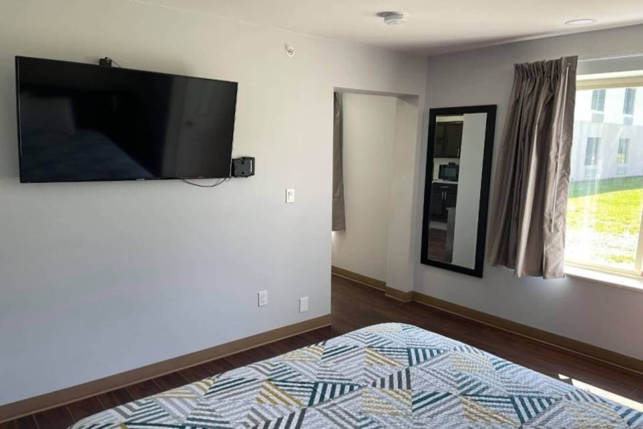 A room with a flat-screen TV mounted on the wall, a patterned rug on the floor, and a window with curtains.