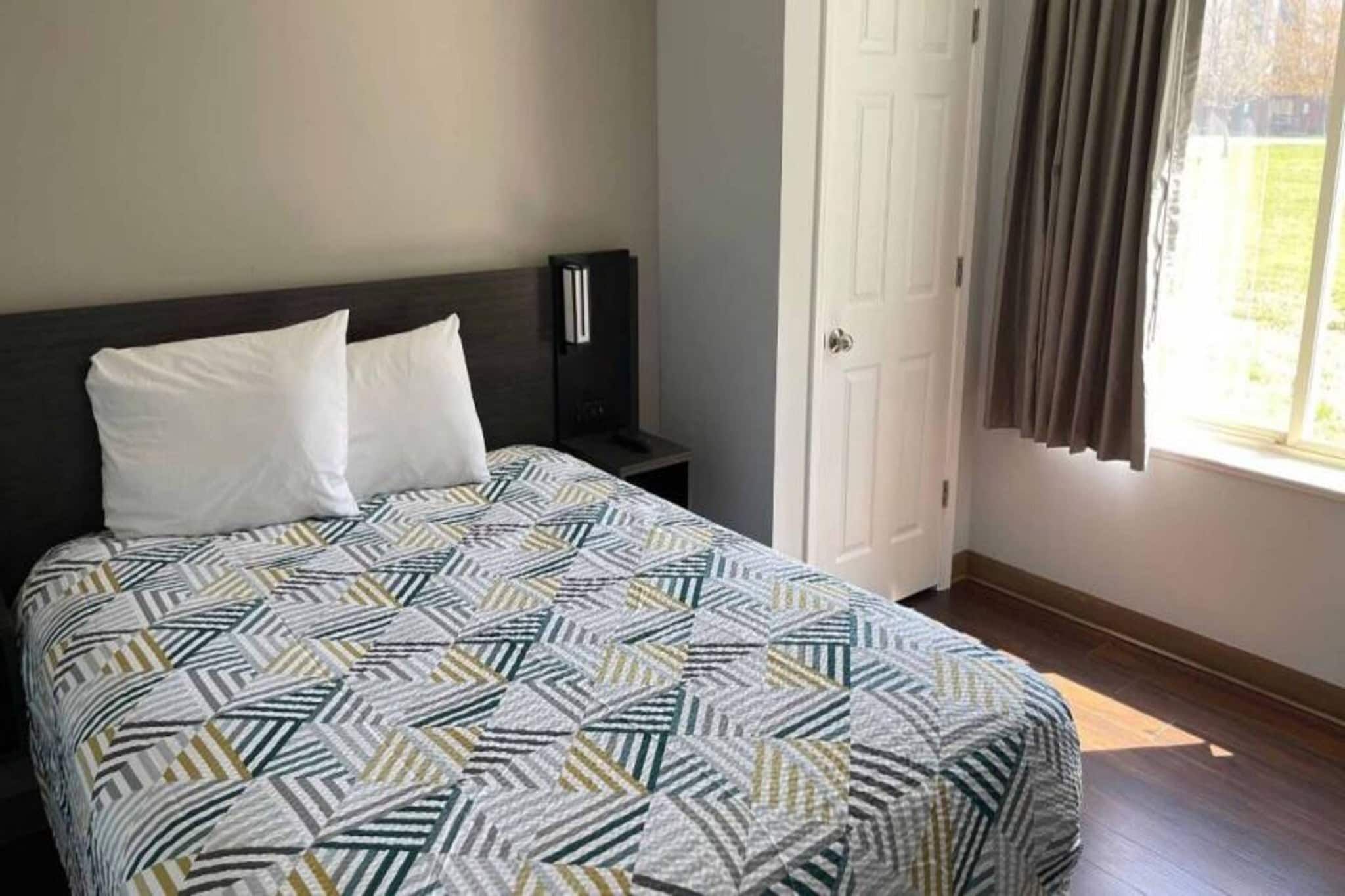 Una cama con una colcha estampada, una mesita de noche con una lámpara, una puerta y una ventana con cortinas.