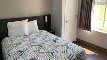 Una cama con una colcha estampada, una mesita de noche con una lámpara, una puerta y una ventana con cortinas.
