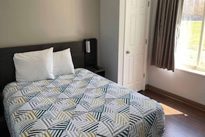 A bed with a patterned bedspread, a bedside table with a lamp, a door, and a window with curtains. - 1 Queen Bed | Non-Smoking, Sofa-Bed (Peoria IL)