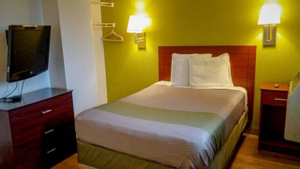 Een hotelkamer met een bed, een televisie op een standaard, een nachtkastje en wandlampen.