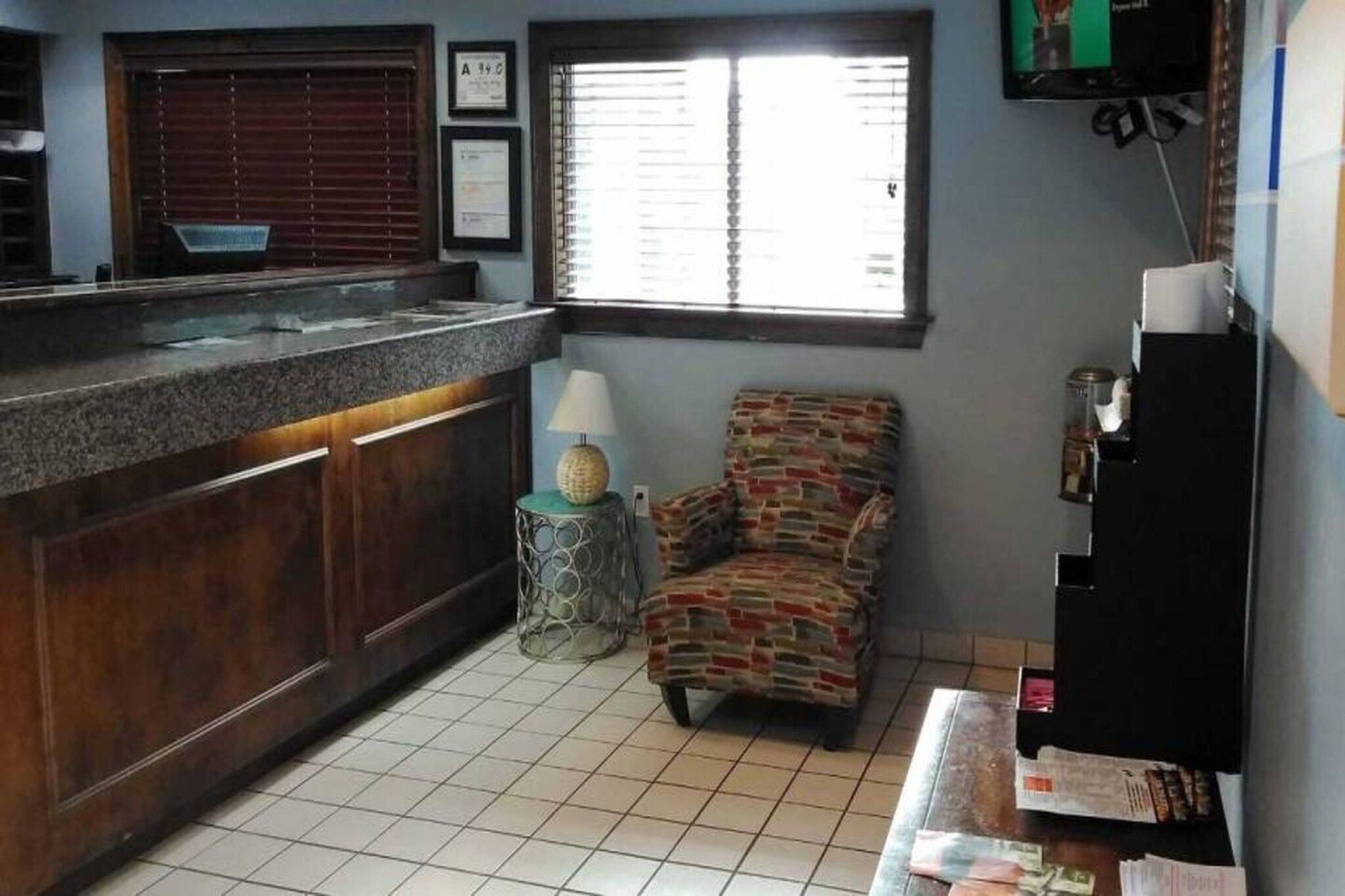 A kitchen with a tiled floor, a wooden counter, a chair, a small table with a lamp, and a window with blinds.