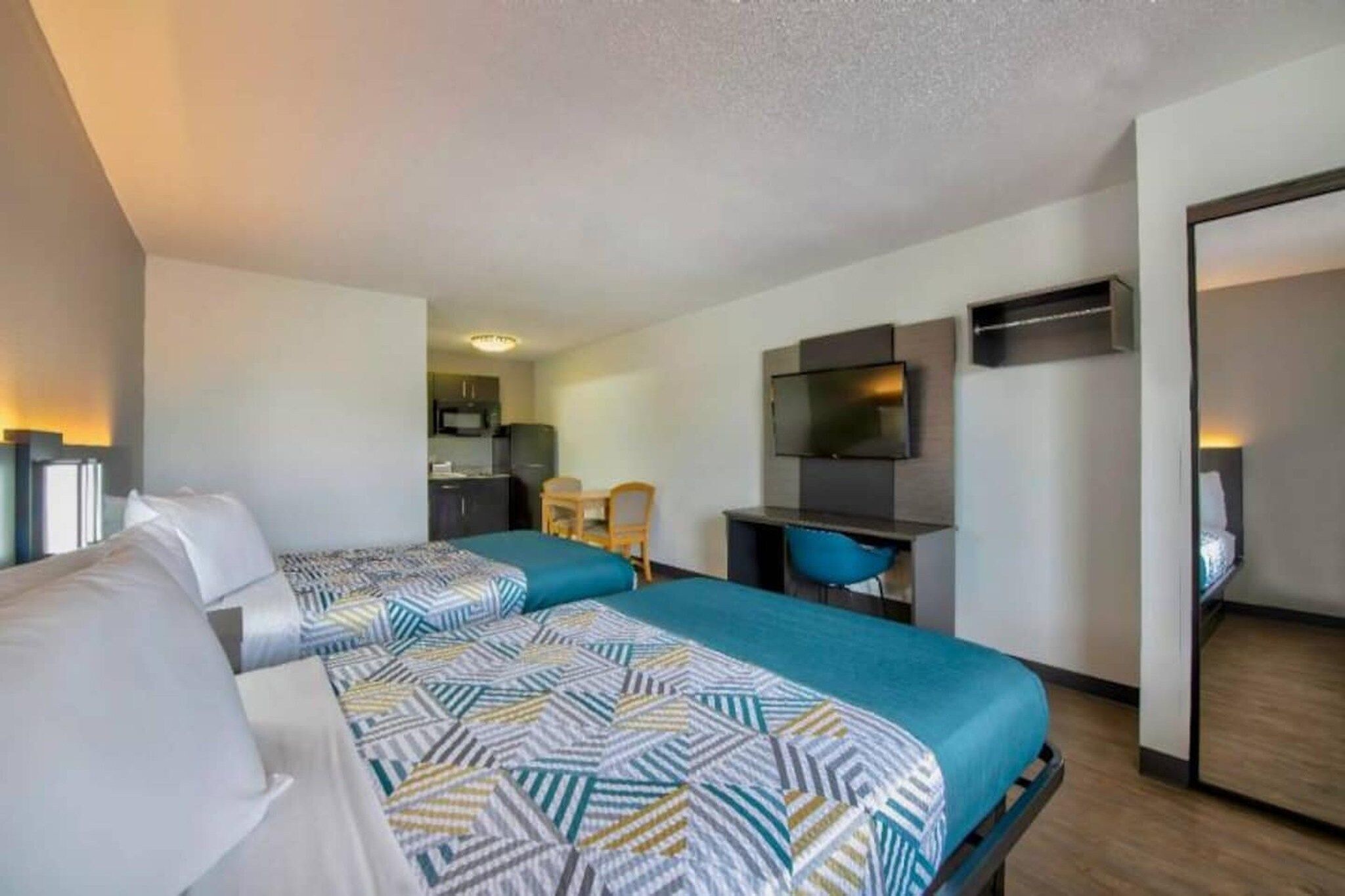 A hotel room with two beds, a desk, a television, and a kitchenette.