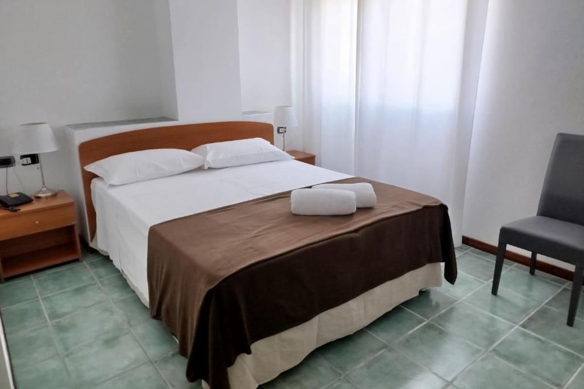 Quarto de hotel com cama, uma cadeira, mesa de cabeceira e abajur.