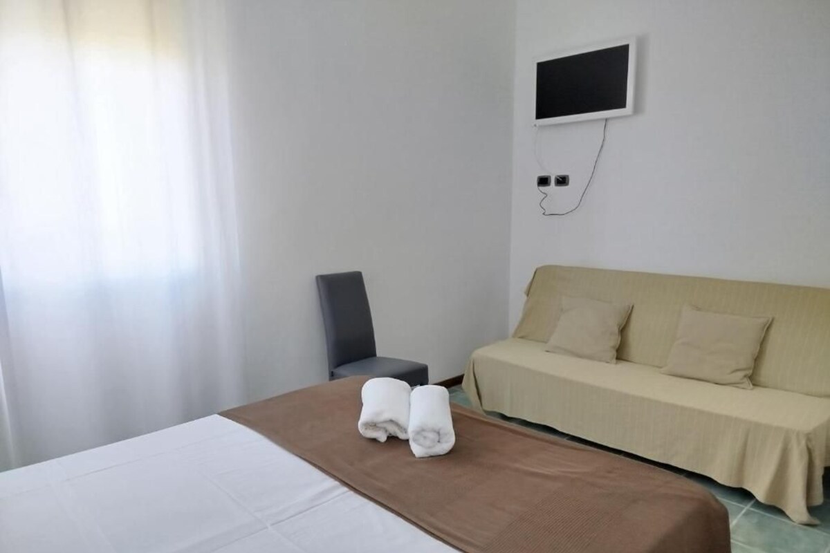 Quarto de hotel com cama, sofá, televisão na parede e janela com cortinas.