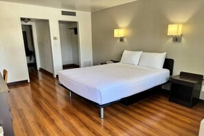 A hotel room with a large bed, bedside tables, a desk, and a chair. There are two wall-mounted lamps and a door leading to another room.