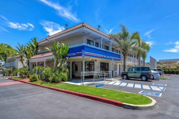 A hotel with a 'WELCOME' sign, a covered entrance, and a parking lot with marked disabled parking spaces. - 1 Queen bed | Los Angeles (Pomona CA)