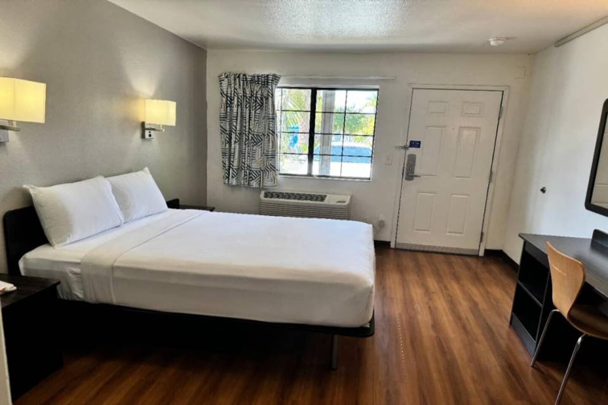 A hotel room with a bed, a desk, a chair, a window with curtains, a door, and a wall-mounted lamp.