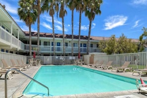 A swimming pool with lounge chairs and a building with balconies and palm trees in the background. - 1 Queen bed | Los Angeles (Pomona CA)