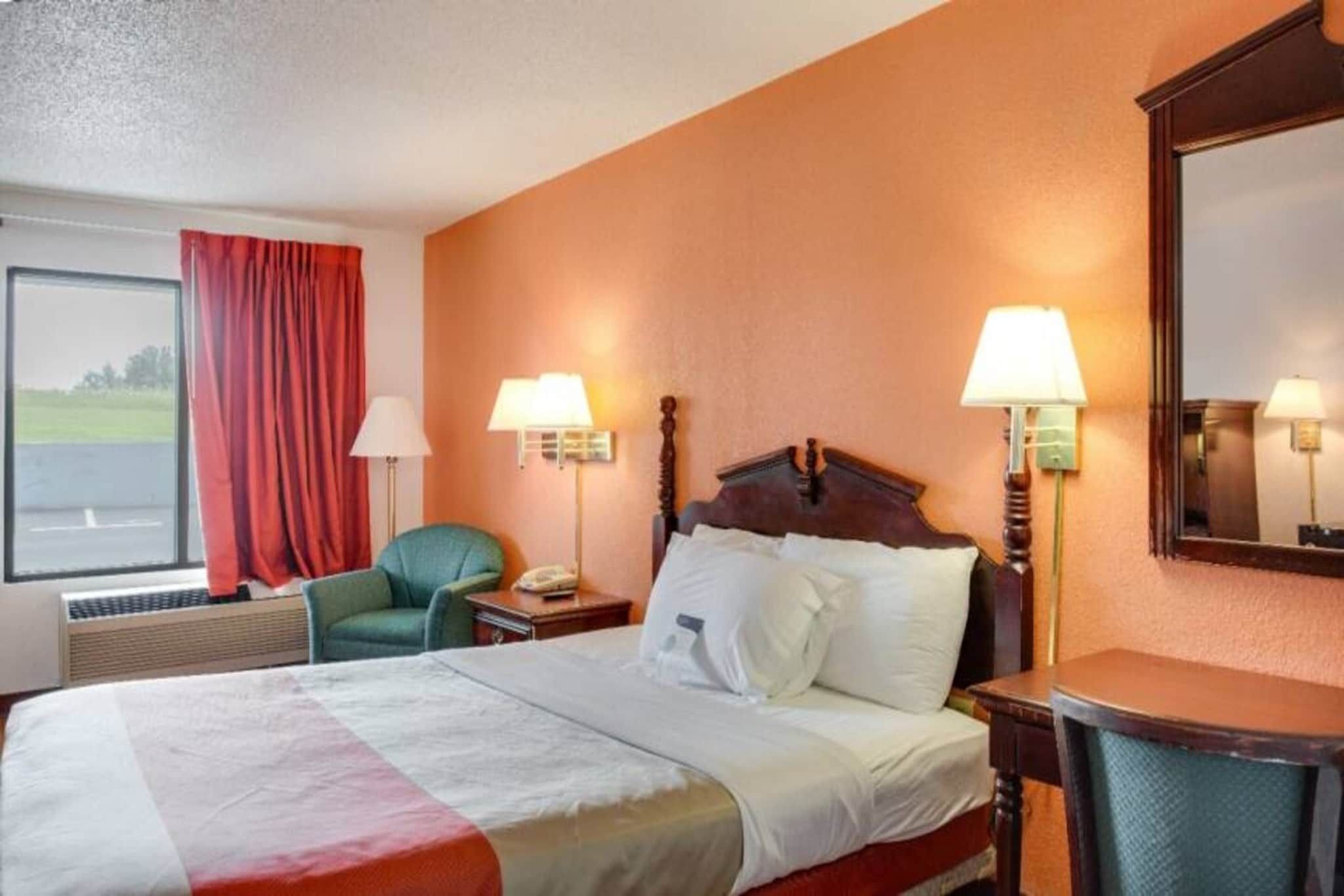 Ein Hotelzimmer mit einem Bett, zwei Nachttischlampen, einem Sessel, einem Fenster mit Vorhängen und Blick auf eine Straße.