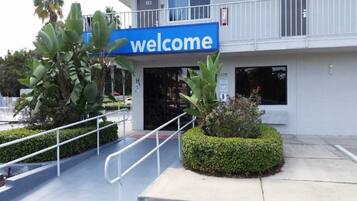 Entrada de edifício com placa de 'welcome', escada e pequena área ajardinada com plantas.