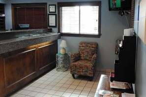 Eine kleine Küche mit gefliestem Boden, einer Holz Arbeitsplatte, einem gemusterten Sessel, einem kleinen Tisch mit Lampe und einem an der Wand befestigten Fernseher.