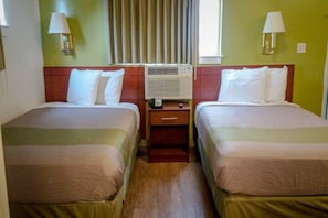 Ein Hotelzimmer mit zwei Betten, einem Nachttisch mit Lampe, einer Klimaanlage und einem Fenster mit Vorhängen.