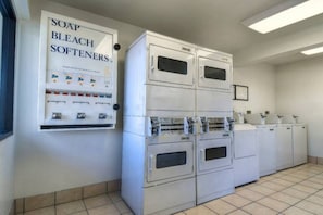 Eine Wäscherei mit gestapelten Waschmaschinen und Trocknern, einem Spender für Waschmittel, Bleichmittel und Weichspüler sowie einem gefliesten Boden.