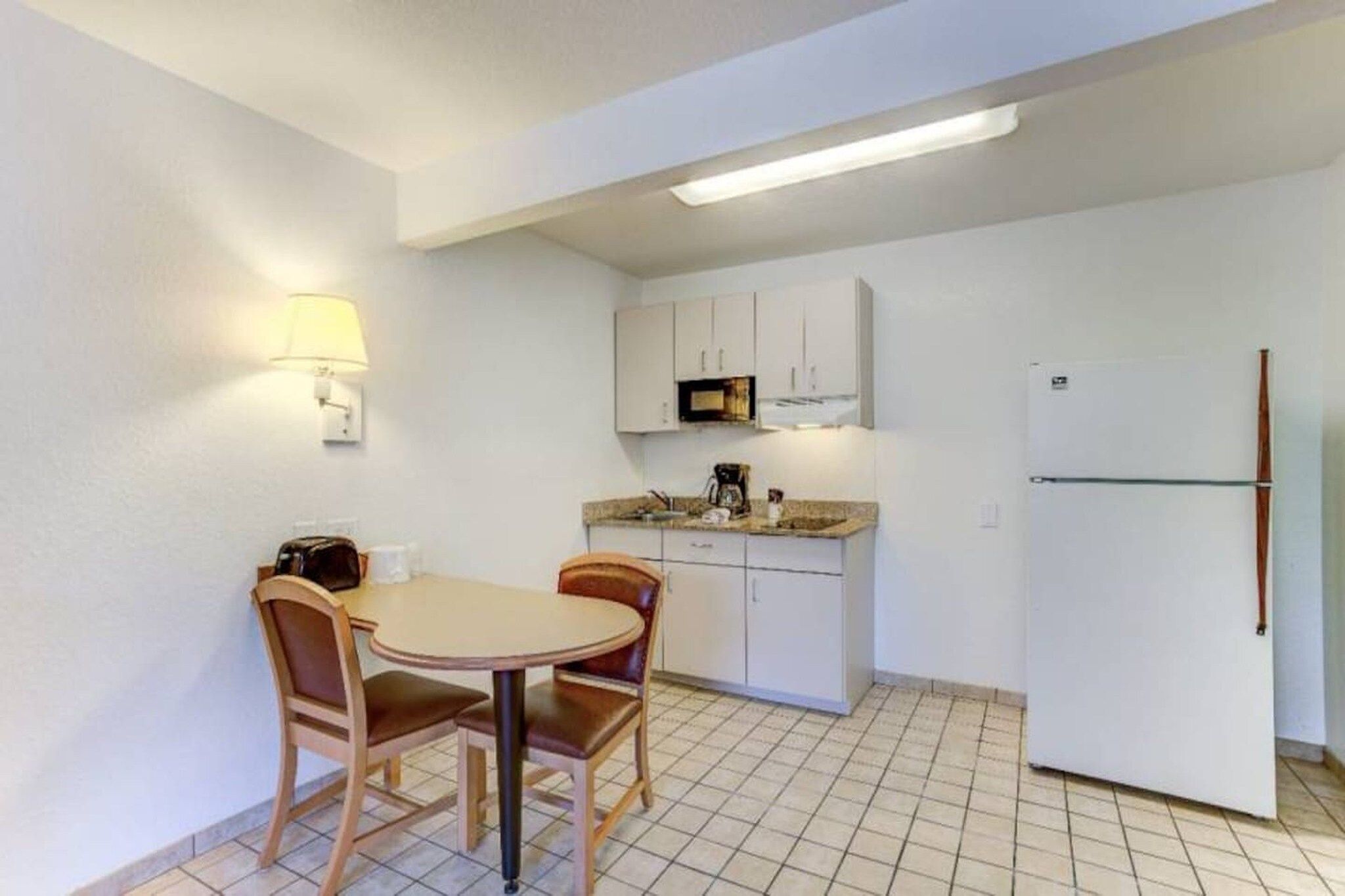 A compact kitchen with a dining table and chairs, a microwave, a refrigerator, and a wall-mounted lamp.