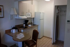 Eine kompakte Küchenzeile mit Mikrowelle, Kaffeemaschine und Kühlschrank. Es gibt eine kleine Essecke mit einem Stuhl und Tisch, sowie einen Fliesenboden.