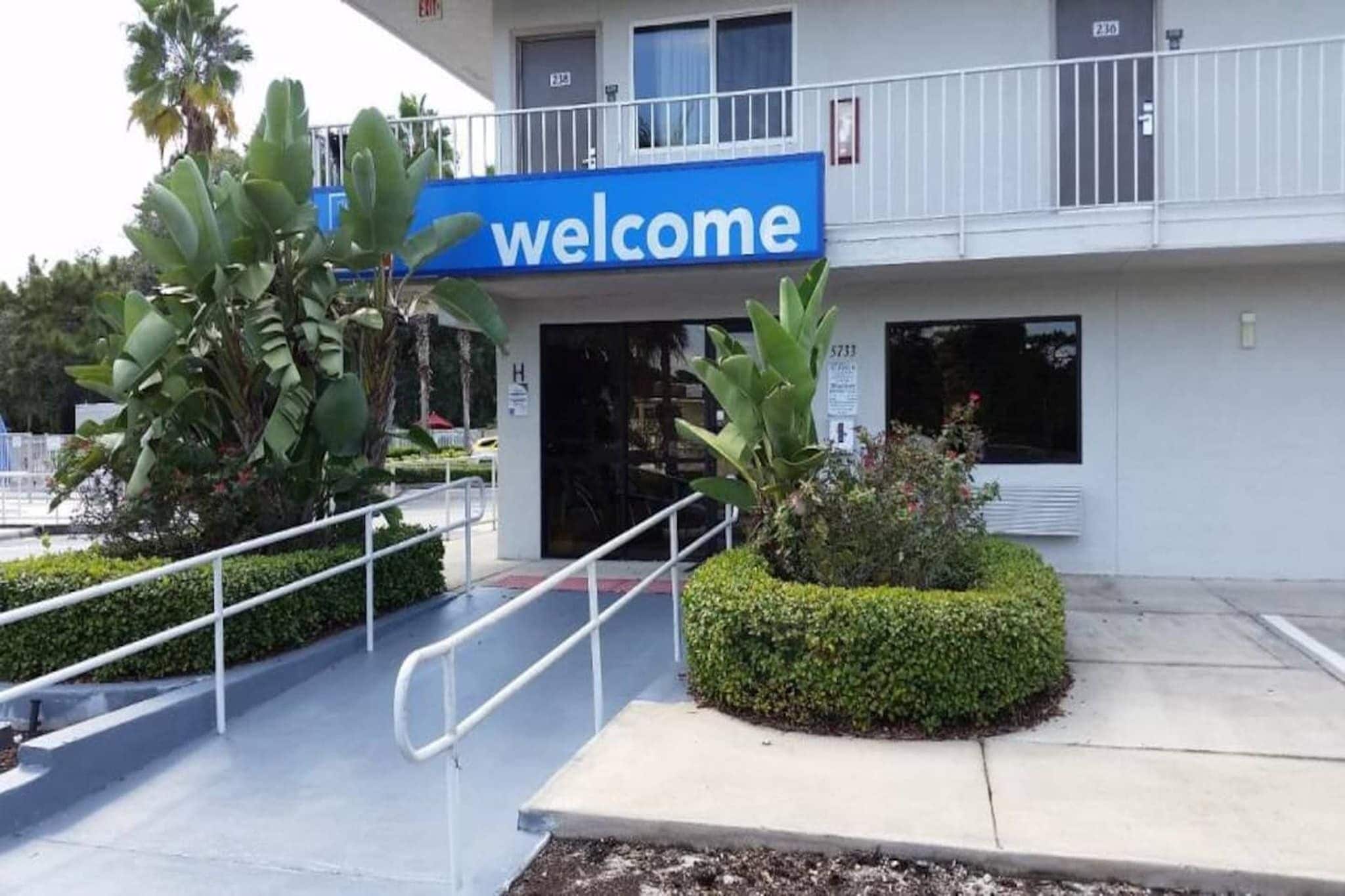 Een gebouwtoegang met een 'welcome'-bord, een trap en een klein aangelegde tuin met planten.